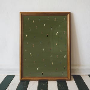 May include: A framed art print featuring a green background with numerous tiny figures, possibly golfers, scattered across the surface. The figures are dressed in various colors. The frame is a warm brown color.