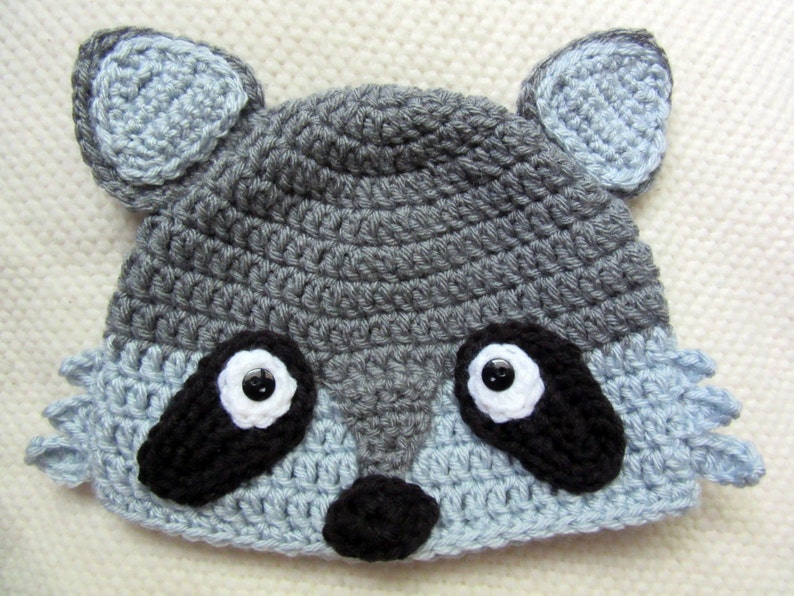 May include: A gray and blue crocheted raccoon hat with black eyes and a black nose.