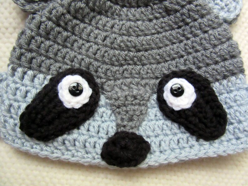 May include: A gray and blue crocheted hat with black crocheted raccoon features, including eyes, nose, and ears.