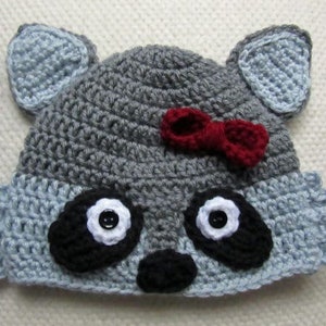 May include: A gray and blue crocheted raccoon hat with black eyes, a black nose, and a red bow.