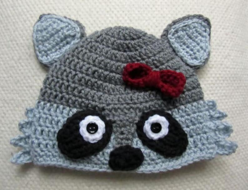 May include: A gray and blue crocheted raccoon hat with black eyes, a black nose, and a red bow.