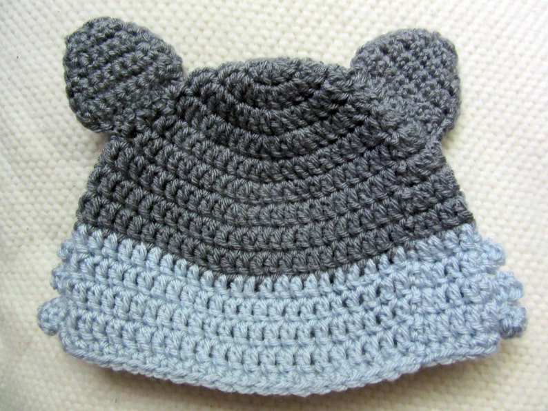 May include: A gray and light blue crocheted hat with two ear flaps. The hat is made with a simple stitch pattern.