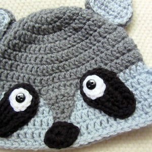 May include: A gray and blue crocheted raccoon hat with black eyes and a black nose.