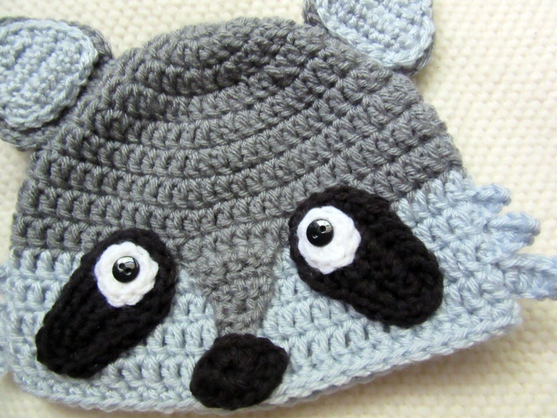 May include: A gray and blue crocheted raccoon hat with black eyes and a black nose.