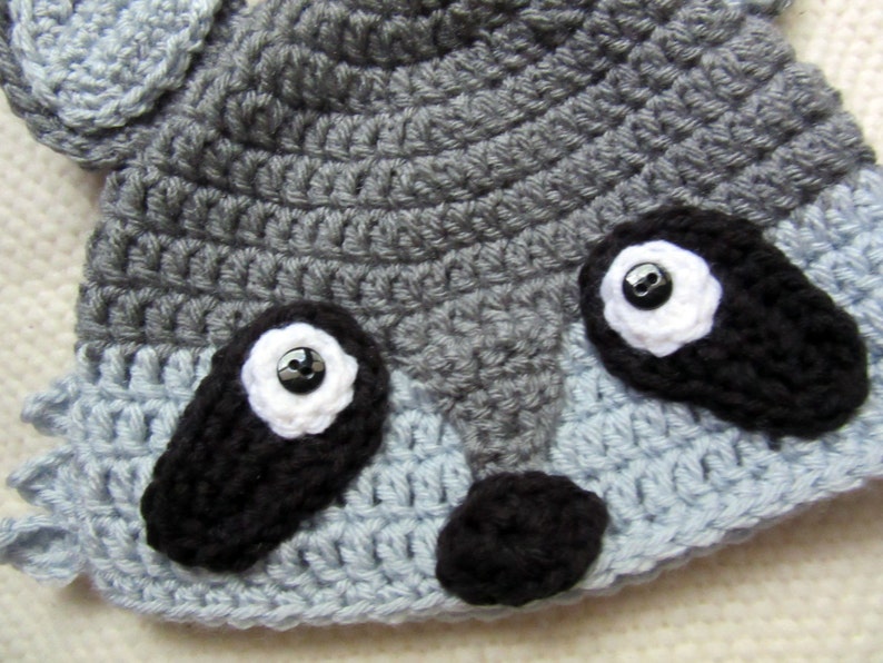 May include: A gray and blue crocheted raccoon hat with black felt eyes and a black felt nose.
