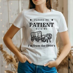 Op de afbeelding: Wit T-shirt met een zwart grafisch ontwerp. De grafiek toont een wagen die door twee paarden wordt getrokken en de tekst "Wees geduldig met me, ik kom uit de jaren 1900".
