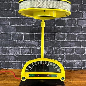 Puede incluir: Una lámpara amarilla única con una base de tablero de instrumentos de coche antiguo y una pantalla de filtro de aire. El tablero muestra un velocímetro, indicadores de combustible, aceite y temperatura. La lámpara está montada sobre una base de madera oscura.