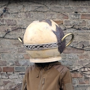 May include: A person wearing a brown fleece jacket and matching sweatpants with a large, round, cream-colored ceramic pot on their head. The pot has a brown glaze and a gold handle. The person is standing in front of a brick wall with snow and leaves on the ground.