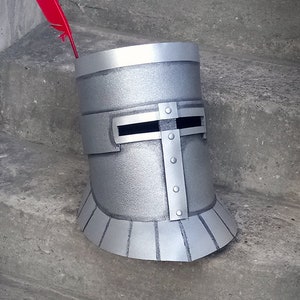 Pode incluir: Capacete de cavaleiro prateado com uma pena vermelha, com viseira e detalhes rebitados. O capacete é cilíndrico com uma base alargada. O logotipo Etsy Black Keep é visível na parte inferior.