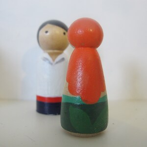 May include: A pair of wooden peg dolls, one with a white shirt and red and blue trim, the other with an orange top and green leaves.