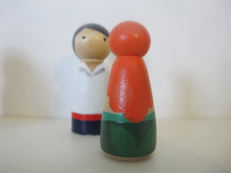 May include: A pair of wooden peg dolls, one with a white shirt and red and blue trim, the other with an orange top and green leaves.
