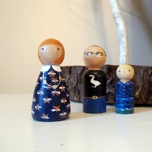 Può includere: Tre bambole di legno con perni, una con i capelli rossi e un vestito a motivi blu e arancioni, una con i capelli castani e una camicia nera con un disegno di un uccello bianco e una con i capelli biondi e un vestito blu.