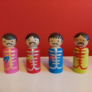 May include: Four wooden peg dolls painted to resemble the Beatles. Each doll has a different coloured outfit: pink, red, blue, and yellow. The dolls have painted facial features, including moustaches.