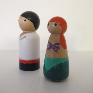 May include: A pair of wooden peg dolls, one with black hair and a red, white, and blue outfit, and the other with red hair and a green and purple outfit.