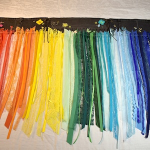 May include: A rainbow-colored fabric banner with a black fabric backing. The banner is decorated with colorful ribbons and lace, and has a row of small embroidered flowers across the top.