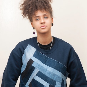 Puede incluir: Una sudadera azul oscuro con un diseño de patchwork hecho de diferentes tonos de mezclilla azul. La sudadera tiene cuello redondo y mangas largas.