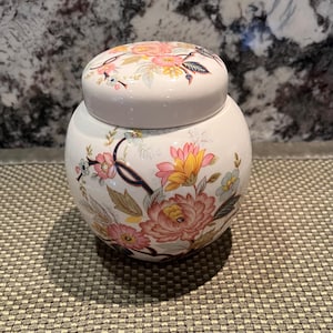 May include: A round white ceramic jar with a matching lid, featuring a colorful floral pattern with pink, yellow, and blue flowers, green leaves, and dark blue stems. The surface of the jar is smooth and glossy, showcasing a detailed botanical design.