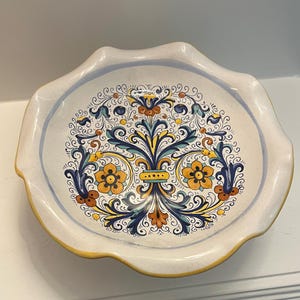May include: A decorative ceramic plate with a scalloped edge. The plate features a central floral design in yellow, blue, and orange against a cream background. The rim has a blue and yellow border.