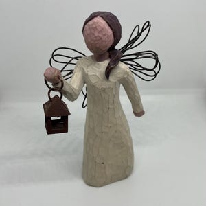 May include: A cream-coloured angel figurine, holding a small brown lantern. The angel has dark hair and wire wings. The figurine is carved with a textured finish, giving it a rustic or folk art aesthetic. The angel's dress is long-sleeved.