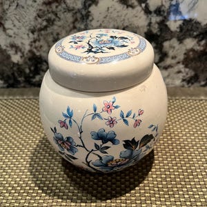 May include: A round, cream-colored ceramic jar with a matching lid, featuring a hand-painted floral design in shades of blue and pink. The pattern includes delicate branches, leaves, and blossoms wrapping around the body and the lid.