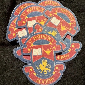 May include: A stack of St. Matthew's Academy stickers. Each sticker features a crest design with the text "ST. MATTHEW'S ACADEMY" in red and gold. The crest includes a book, lamp, and a yellow emblem on a blue background.
