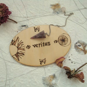 May include: A wooden pendulum board with a sun and moon design. The board has the words "yes", "no", and "veritas" written on it. A pink crystal pendulum hangs from a silver chain.