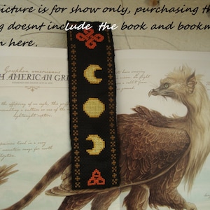 Puede incluir: Un marcapáginas negro y dorado con un patrón repetido de lunas y un símbolo de triskel. El marcapáginas está sobre un libro con una imagen de una criatura mítica. El texto del libro dice "Gryphon americanus NORTH AMERICAN GRIFFIN", "Aunque descendiente de un águila, este grifo se parecía a un buitre en el uso de las rapaces ligeras." y "Este espécimen vivió en una cordillera muy remota para la profundidad de la virilización."