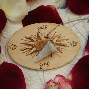 Könnte beinhalten: Ein Holz-Pendelbrett mit den Worten "ja", "nein" und einem Sonnenmotiv. Ein weißes Kristallpendel hängt an einer silbernen Kette.