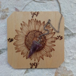 Könnte beinhalten: Ein Holz-Pendelbrett mit einem Sonnenblumenmuster und den Worten "ja" und "nein" darauf. Ein violettes Kristallpendel hängt an einer silbernen Kette.