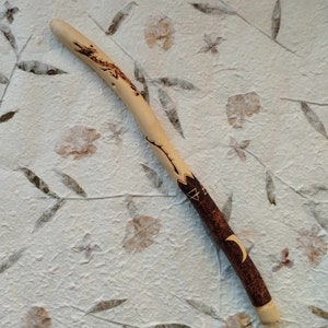 Może przedstawiać: Drewniana różdżka z brązowo-czarnym wzorem. Różdżka ma wyrzeźbiony półksiężyc i symbol trójkąta.