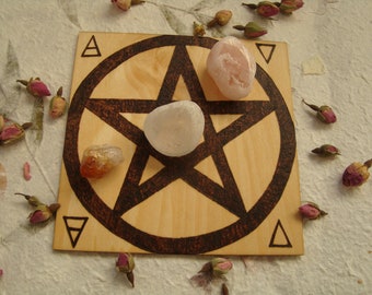 May include: A wooden board with a pentagram and circle design burned into the surface. There are three crystals on the board: a clear quartz crystal in the center, a pink rose quartz crystal to the right, and a clear citrine crystal to the left. The board has four triangles burned into the corners.