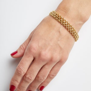 May include: A gold chain bracelet with a delicate, interwoven design. The bracelet is worn on a wrist with red nail polish.