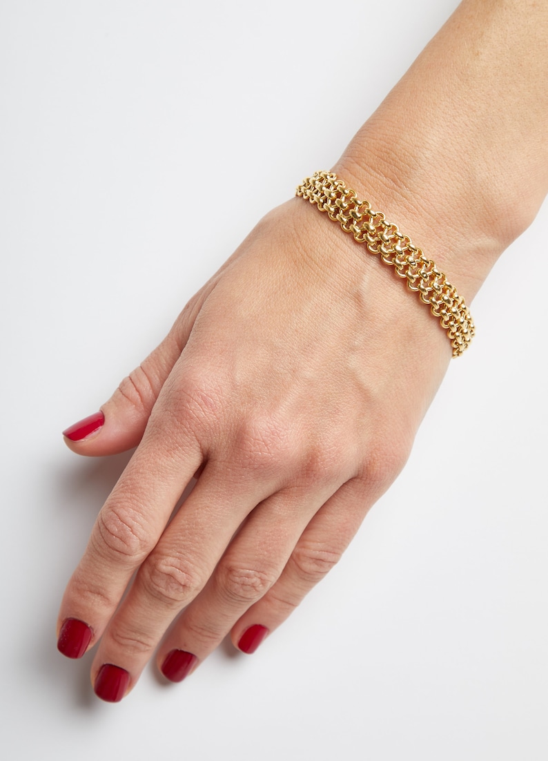 May include: A gold chain bracelet with a delicate, interwoven design. The bracelet is worn on a wrist with red nail polish.