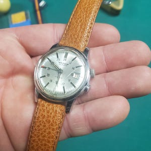 May include: A vintage Waltham Alarm wristwatch with a silver face, date display, and a brown leather strap. The watch has a stainless steel case and a textured leather band. The watch is held in a hand.