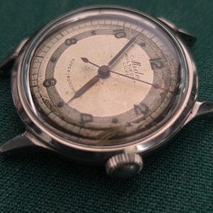 May include: Close-up of a vintage silver wristwatch with a cream-colored face. The watch features black hands, a second hand, and a small crown. The watch face has the words "Mido Multifort Luxe" and "Super-Automatic".