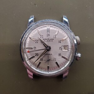 May include: A vintage Waltham-Am alarm watch with a silver face and a stainless steel case. The watch face displays the time, date, and the text "WALTHAM ALARM". The watch has a red arrow hand and two crowns.