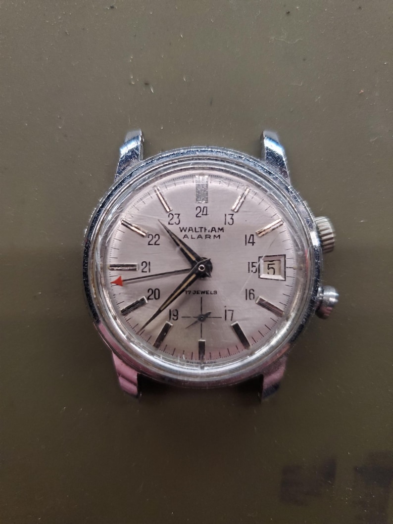 May include: A vintage Waltham-Am alarm watch with a silver face and a stainless steel case. The watch face displays the time, date, and the text "WALTHAM ALARM". The watch has a red arrow hand and two crowns.