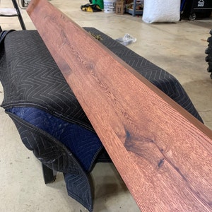 May include: A long, reddish-brown wooden plank with a natural wood grain pattern. The plank is lying on a black and blue moving blanket.