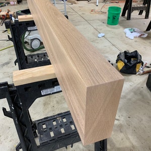 May include: A long, light brown wooden beam resting on two black sawhorses. The beam has a smooth, polished surface and is likely made of oak.