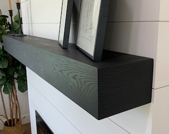 May include: A black wooden mantel shelf with a dark wood grain pattern, mounted above a white fireplace surround with a black fireplace insert. Two black framed pictures are displayed on the shelf.