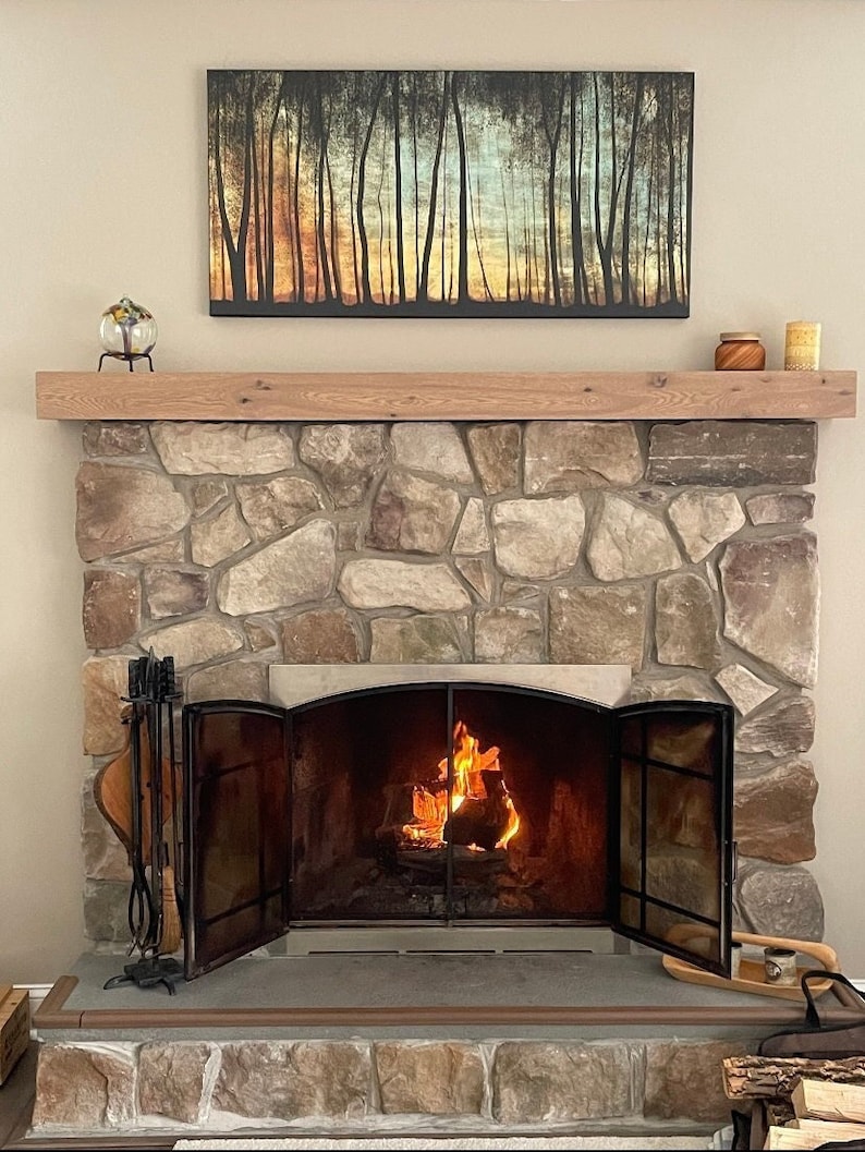 May include: A stone fireplace with a wood-burning fire and a black metal fire screen. The fireplace is decorated with a painting of a forest scene above the mantel.
