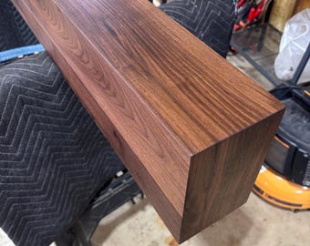 May include: A rectangular wooden beam with a dark brown finish and visible wood grain patterns. The beam features sharp, clean edges and a smooth surface, showcasing the natural texture and deep color variations of the wood material throughout.