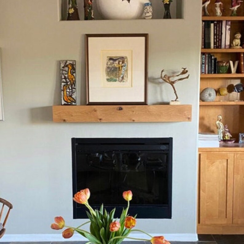May include: A black fireplace with a wood mantel shelf above it. The mantel shelf is decorated with a framed picture, a small sculpture, and a wooden bird figurine. The fireplace is in a room with a light green wall and a wooden bookcase.