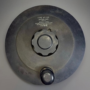May include: A circular antenna reel, model MT-5E, made of dark metal. It has a central gear-like mechanism and a black knob. The text on the reel indicates it was manufactured by Bendix Radio in Baltimore, Maryland, USA.