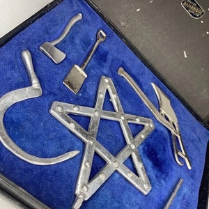 Puede incluir: Un conjunto de herramientas en miniatura en una caja forrada de terciopelo azul. Las herramientas incluyen una pala, un pico, un gancho, una herramienta en forma de estrella y un par de alicates. La caja está etiquetada con "TRADE RAINBRIDGE LONDON".