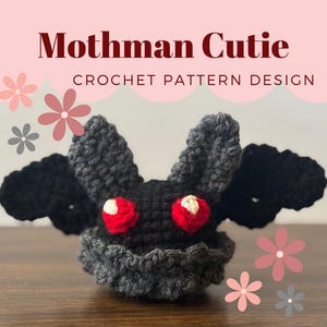 Peut inclure: Un jouet en crochet Mothman Cutie amigurumi avec des ailes noires, des yeux rouges et un corps gris. Le jouet est posé sur une surface en bois.