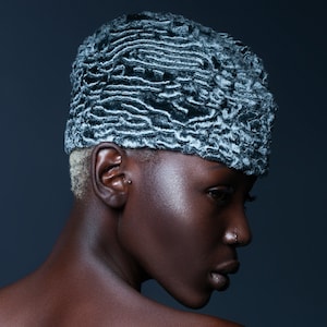 May include: A person wearing a textured, gray and white hat. The hat has a unique, wavy pattern. The person has dark skin and short, blonde hair. The background is a dark blue.