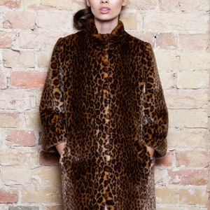 Pu&ograve; includere: Un cappotto lungo con stampa leopardata, colletto alto e tasche. Il cappotto presenta un motivo maculato marrone e nero. La modella &egrave; in piedi davanti a un muro di mattoni. Il cappotto &egrave; lungo fino al ginocchio.