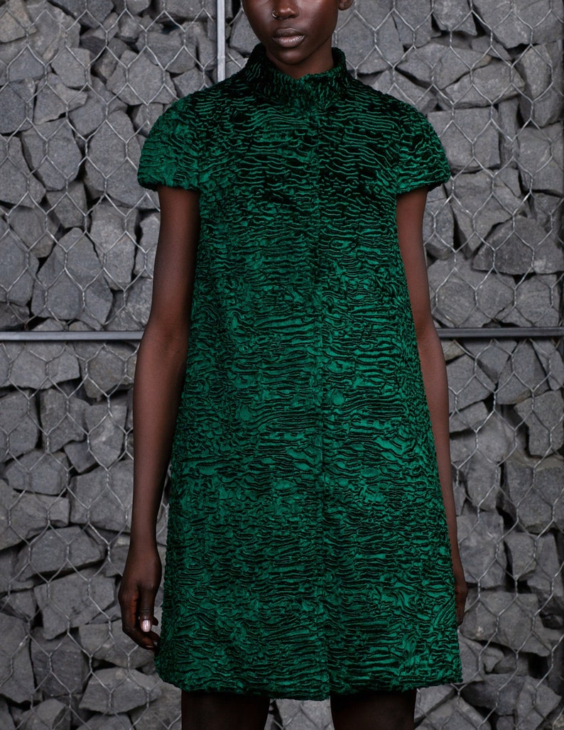 May include: A short-sleeved, emerald green coat with a textured pattern. The coat has a high collar and a knee-length cut. The fabric appears to be a plush material. The background is a gray stone wall.