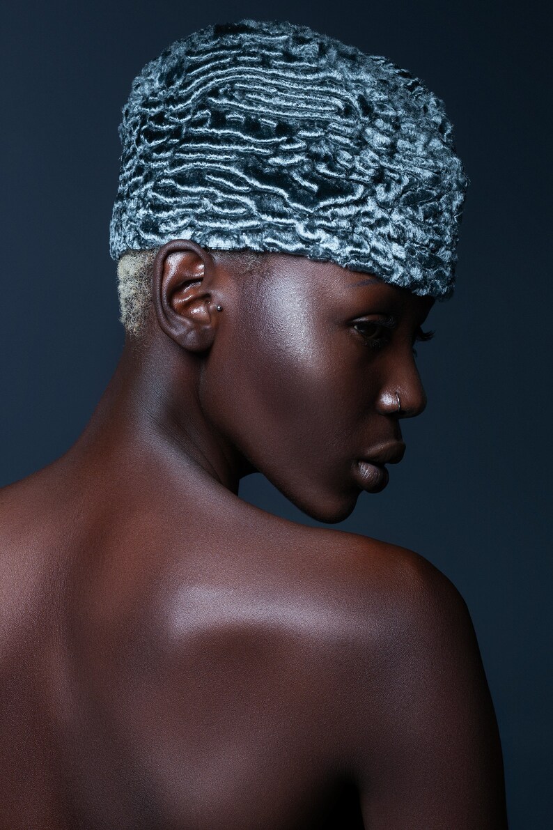 May include: A person wearing a textured, gray and white hat. The hat has a unique, wavy pattern. The person has dark skin and short, blonde hair. The background is a dark blue.
